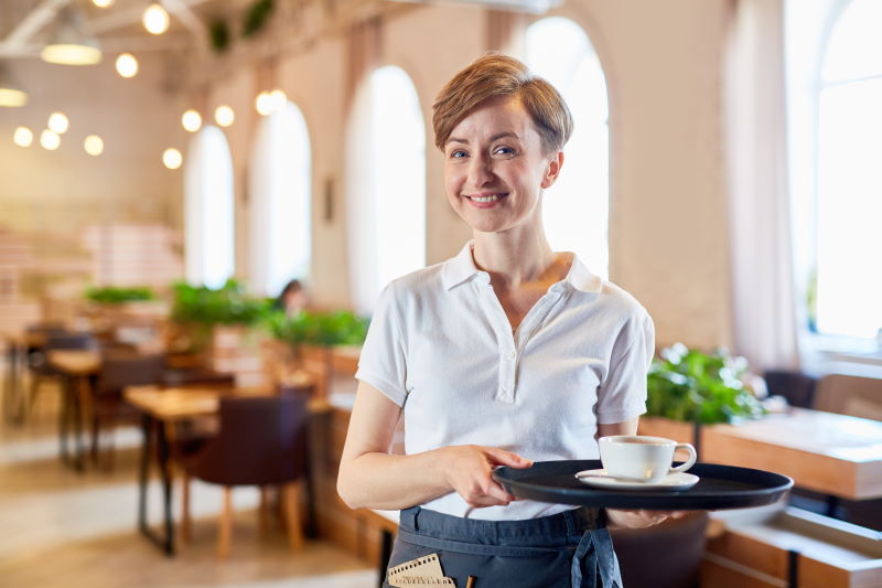 Smiling sesrvant in uniform bringing cup of coffee to visitor of restaurant
